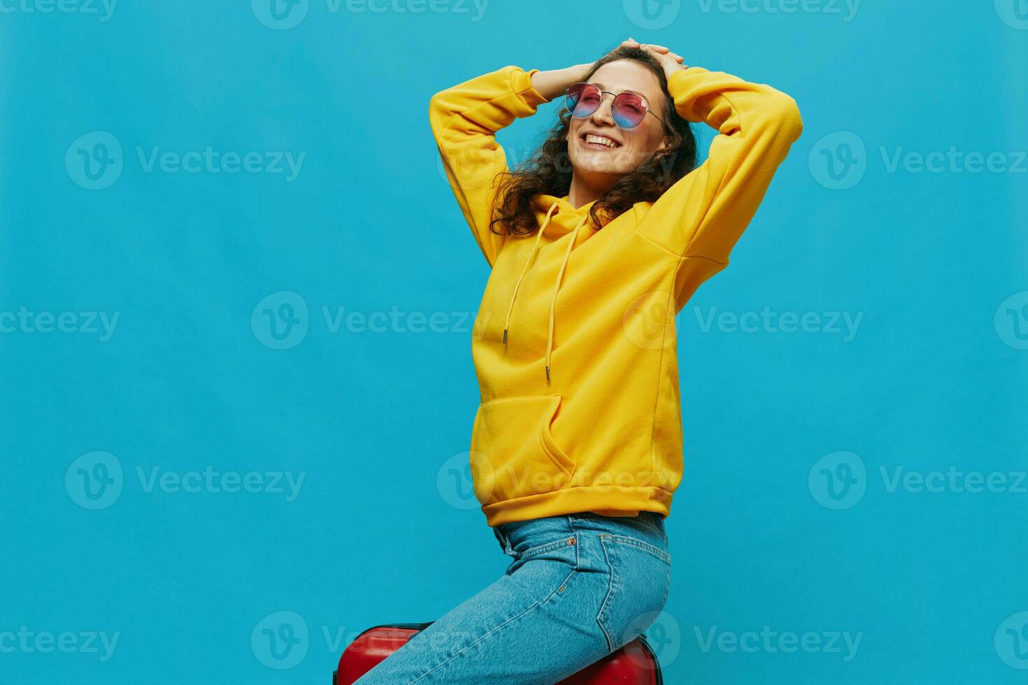 sourire femme séance sur une valise dans une Jaune sweatshirt à capuche, bleu jeans et des lunettes sur une bleu arrière-plan, emballage pour une voyage photo