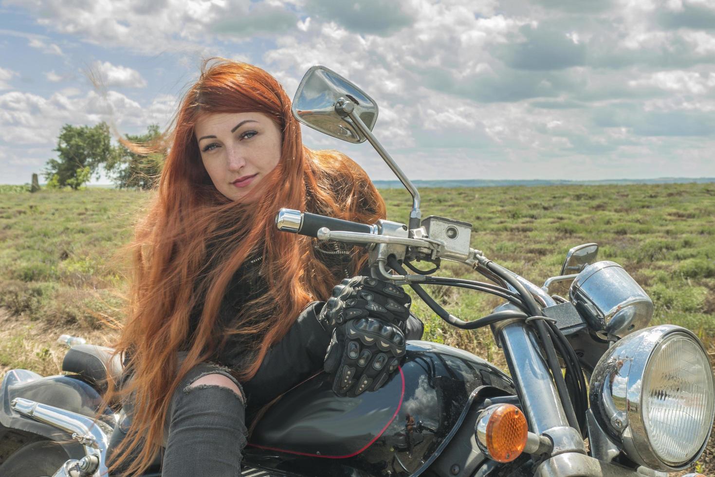 La fille de motard aux cheveux rouges est assise sur un champ de moto de prairie et de nuages photo