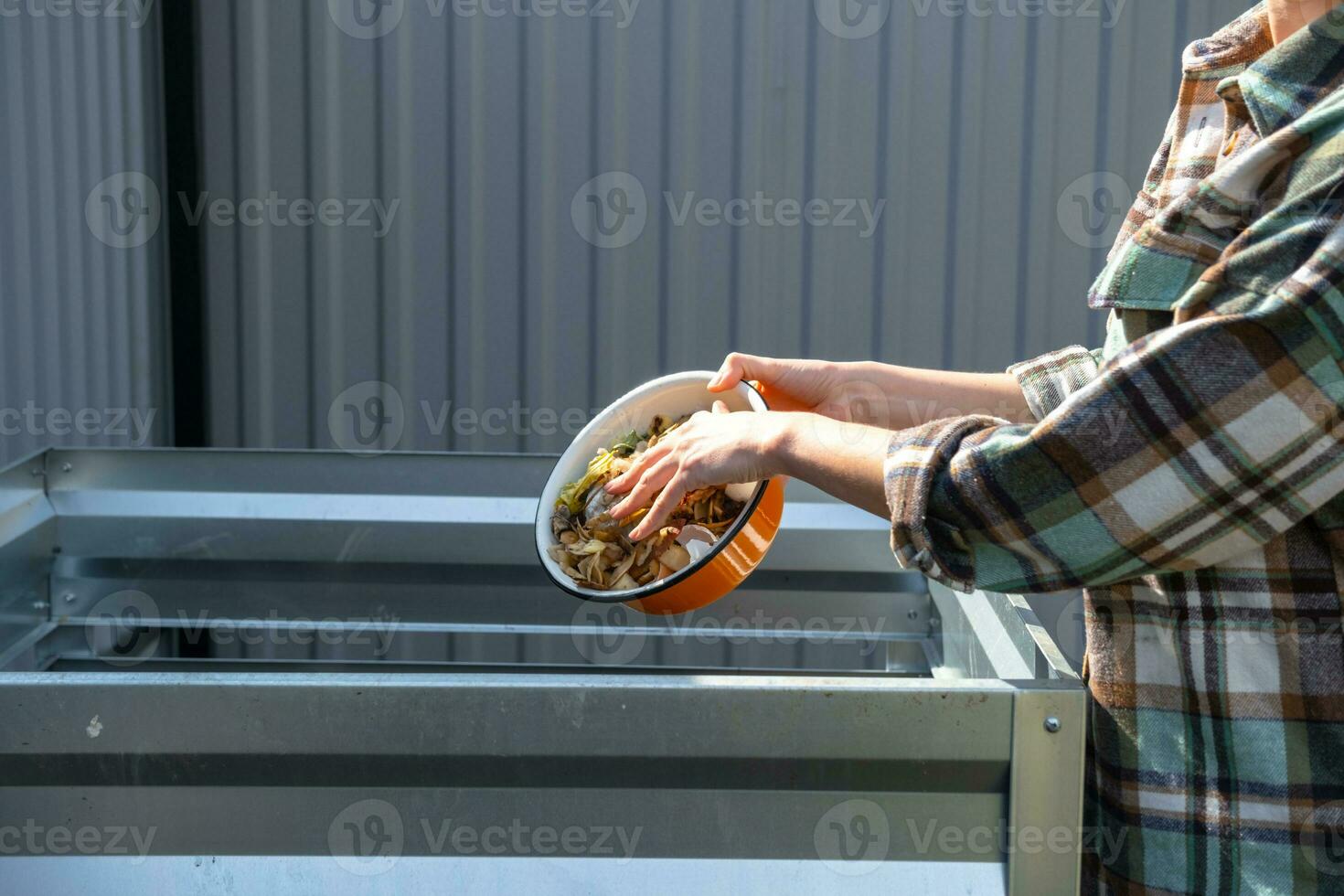 une femme dans une plaid chemise verse nourriture déchets de une bol dans une compost tas de Patate et carotte épluchures. compost boîte fabriqué de métal, respectueux de la nature engrais pour le jardin photo