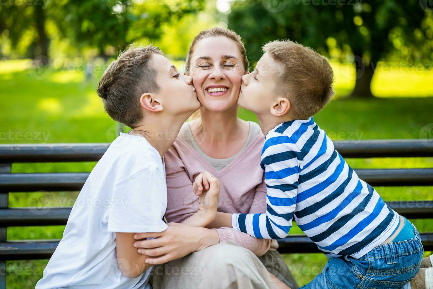 content mère est séance avec sa fils sur banc dans parc. elles ou ils sont ayant amusement ensemble. garçons sont embrasser leur mère. photo