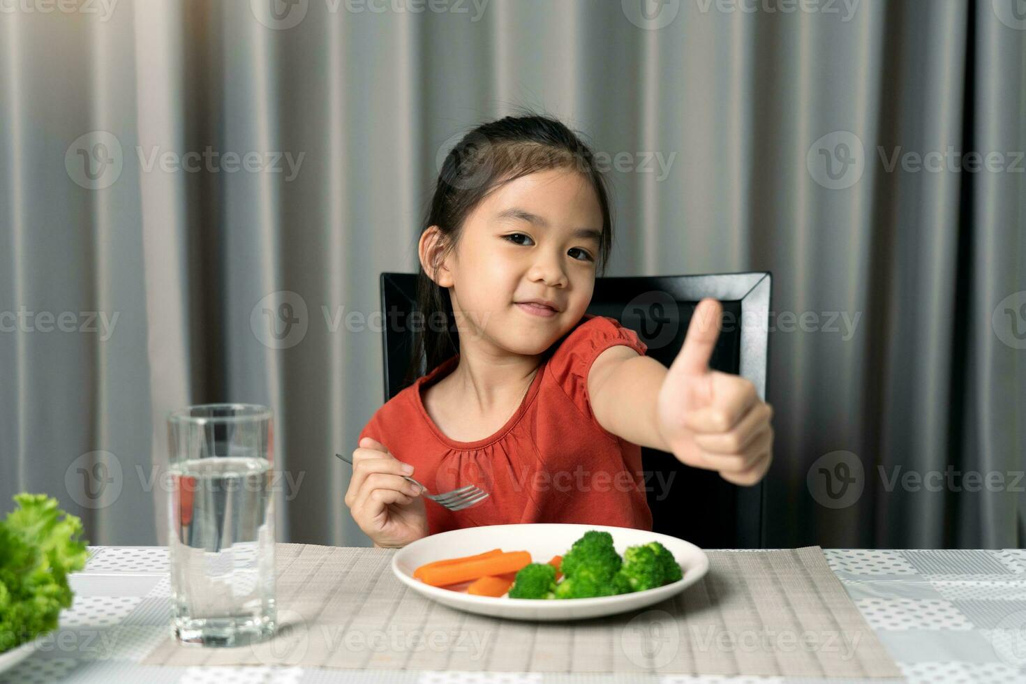 mignonne peu fille montrant pouce montrant en mangeant en bonne santé des légumes. photo