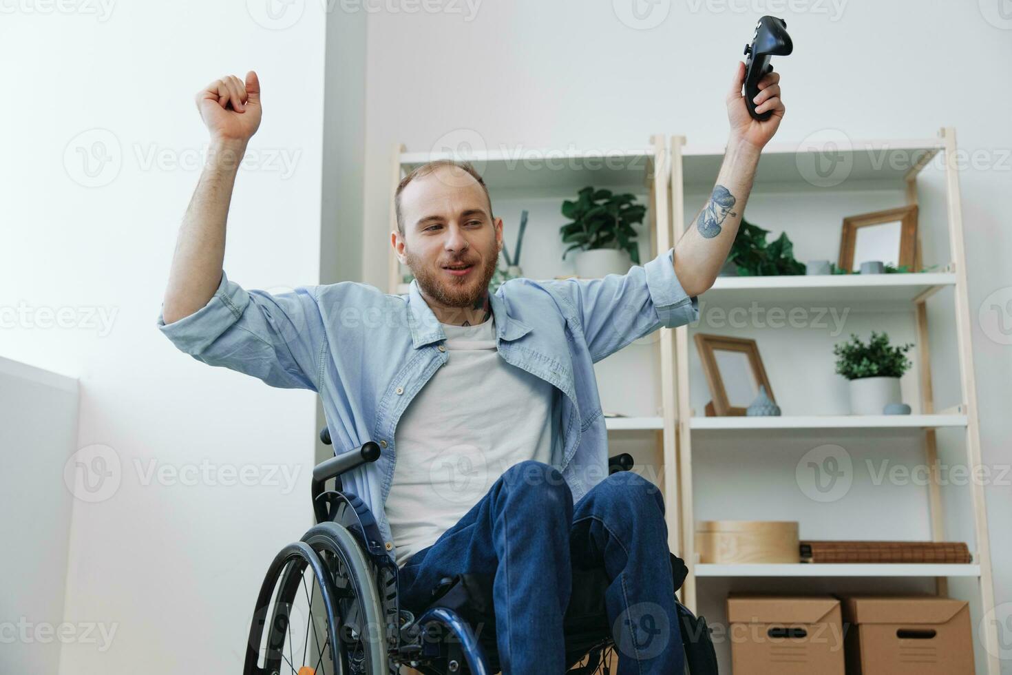 une homme dans une fauteuil roulant joueur pièces Jeux avec une manette dans le sien mains à maison, bonheur, la victoire, mains en haut, copie espace, avec tatouages sur le sien mains, santé concept homme avec handicapées, réel la personne photo