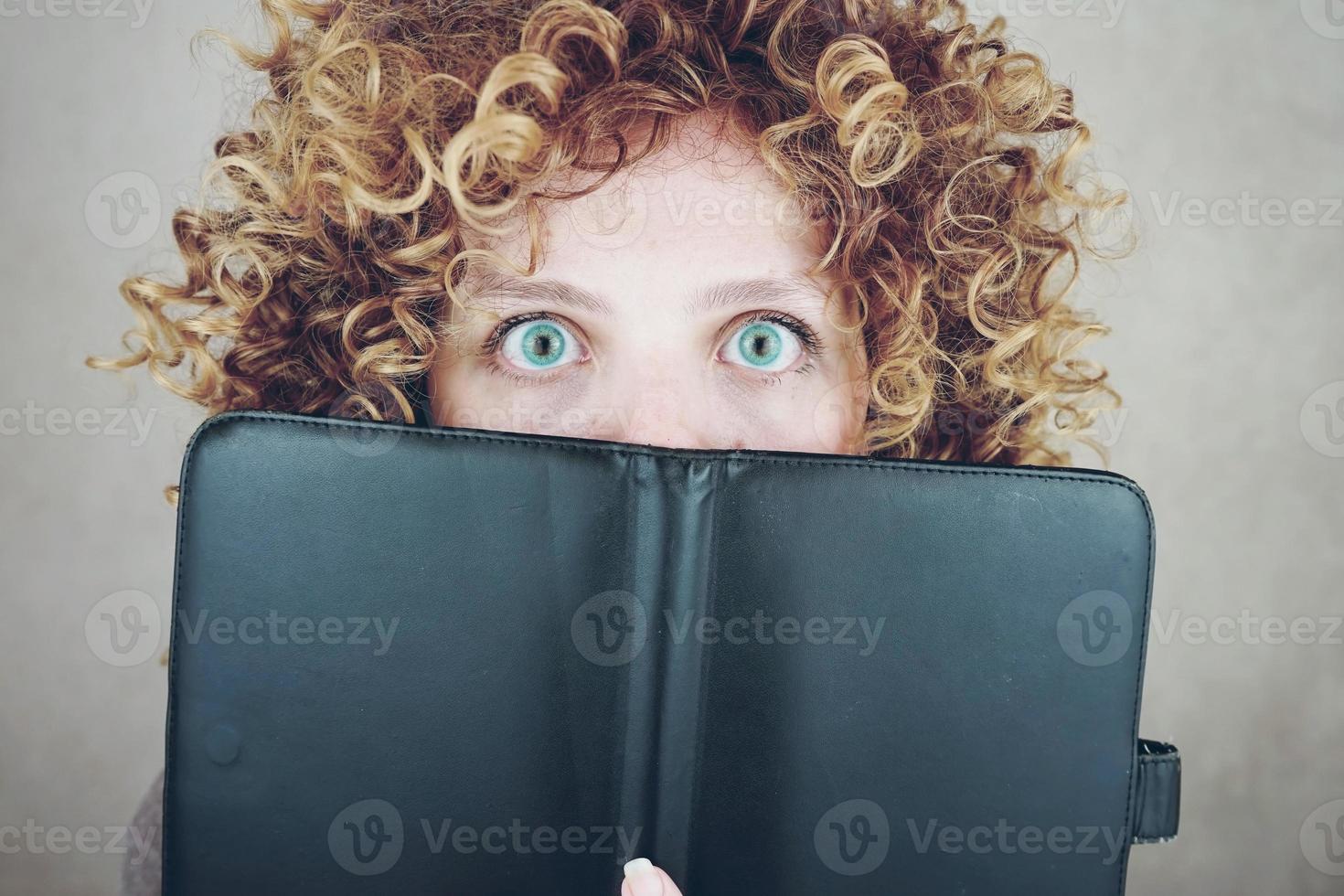 Portrait en gros plan d'une belle et jeune femme drôle aux yeux bleus et aux cheveux blonds bouclés, elle est derrière un agenda ou un ebook et elle est surprise photo