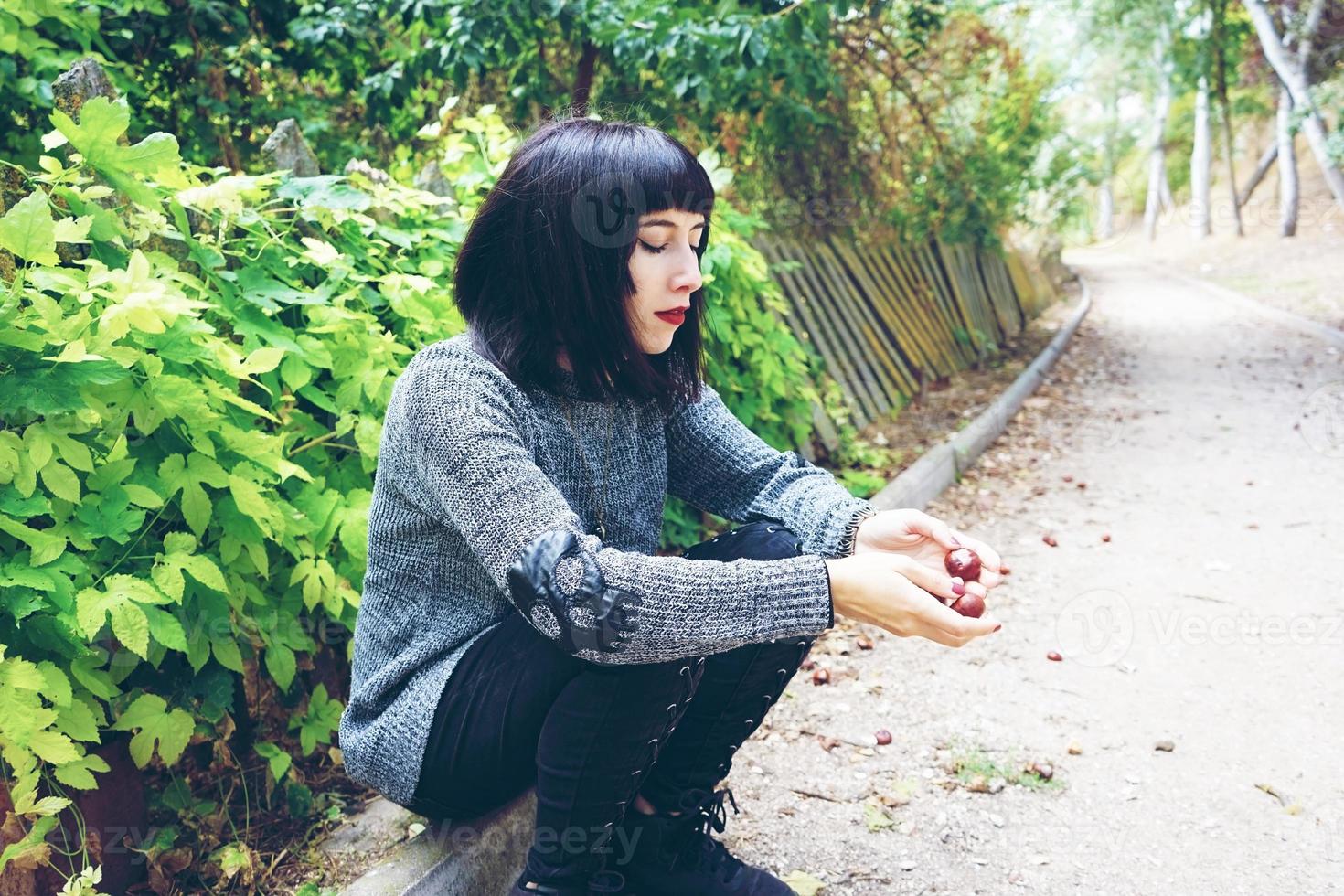 Belle femme brune caucasienne assise sur le sol dans un parc portant des vêtements punk ou gothiques et entourée de feuilles vertes photo