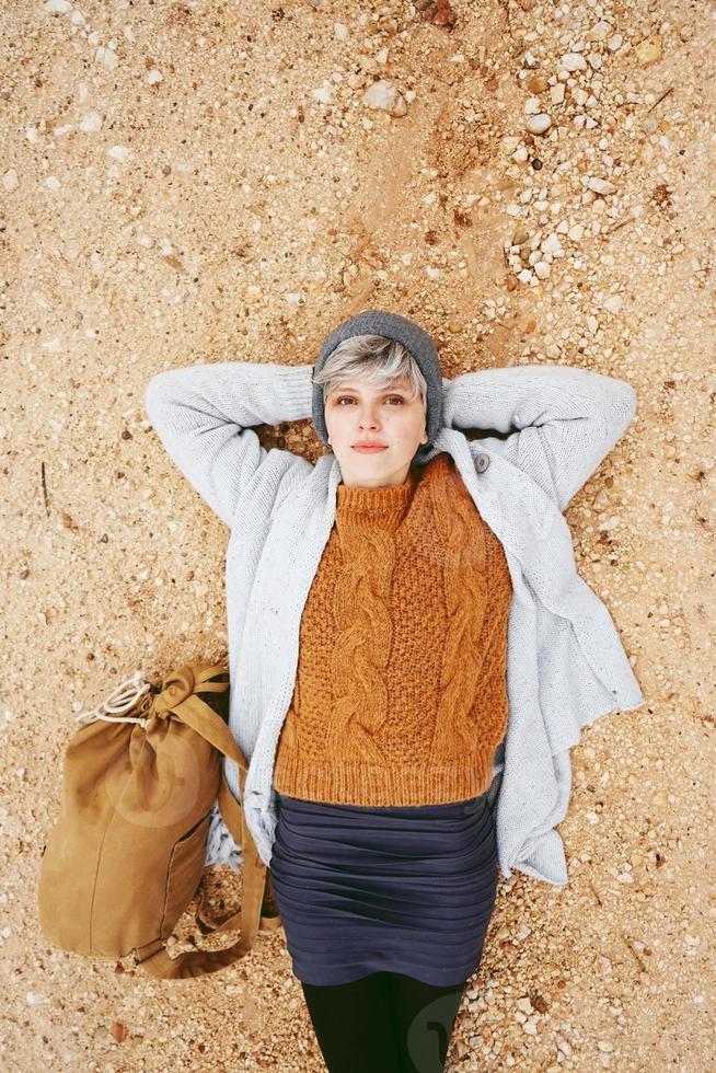 Une jeune femme de race blanche aventurière allongée sur un sol de gravier à côté d'un sac à dos portant un pull en laine et un bonnet en laine gris avec l'orange comme couleur principale photo