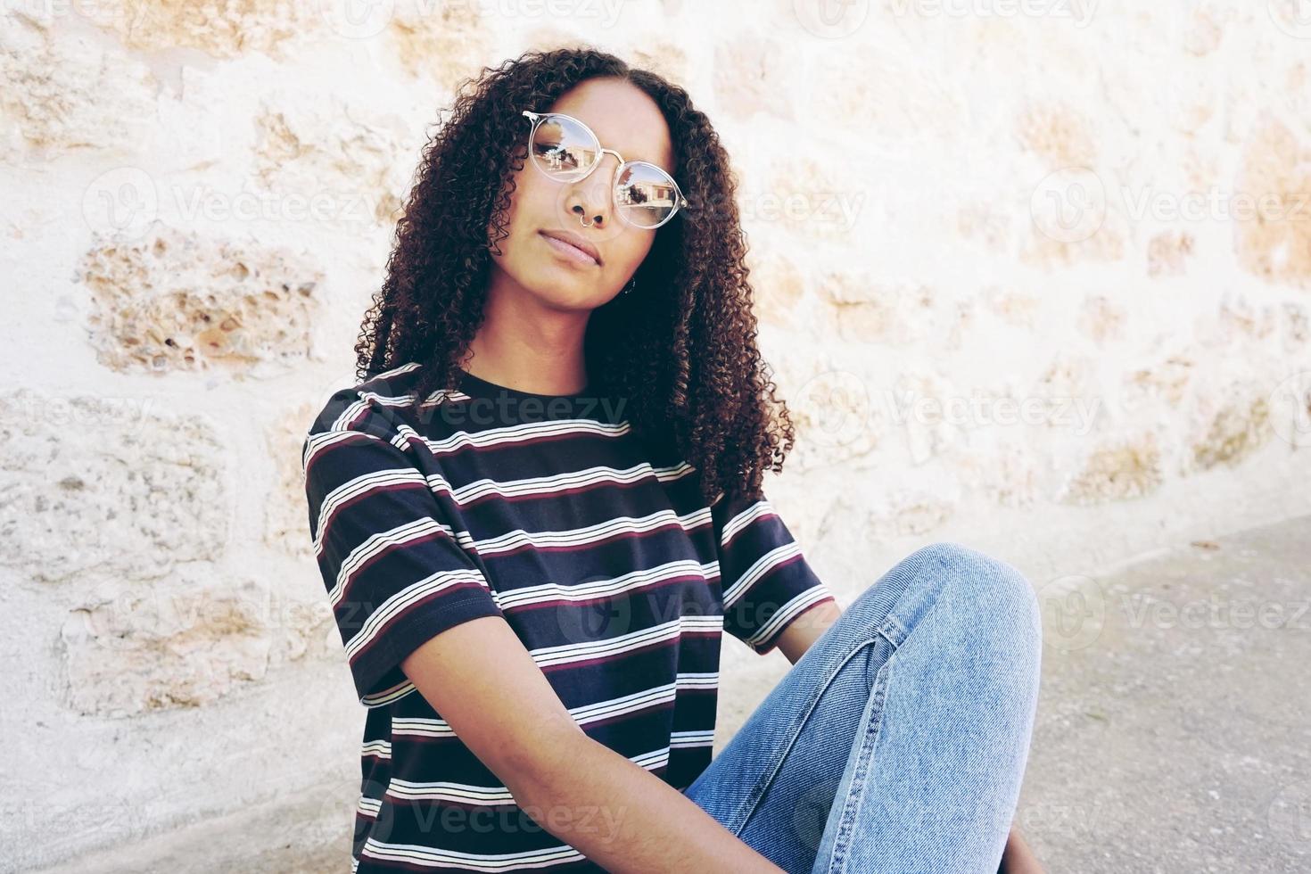 un portrait d'une jeune femme noire sérieuse portant des lunettes, un jean et un t-shirt rayé, assise sur le sol et les cheveux bouclés photo