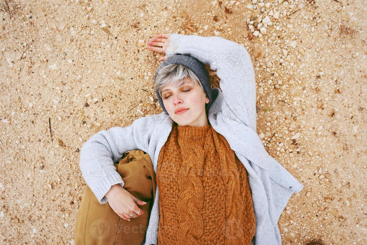 Une jeune femme de race blanche aventurière allongée sur un sol granuleux à côté d'un sac à dos portant un pull en laine et une casquette avec les yeux fermés et l'orange comme couleur principale photo