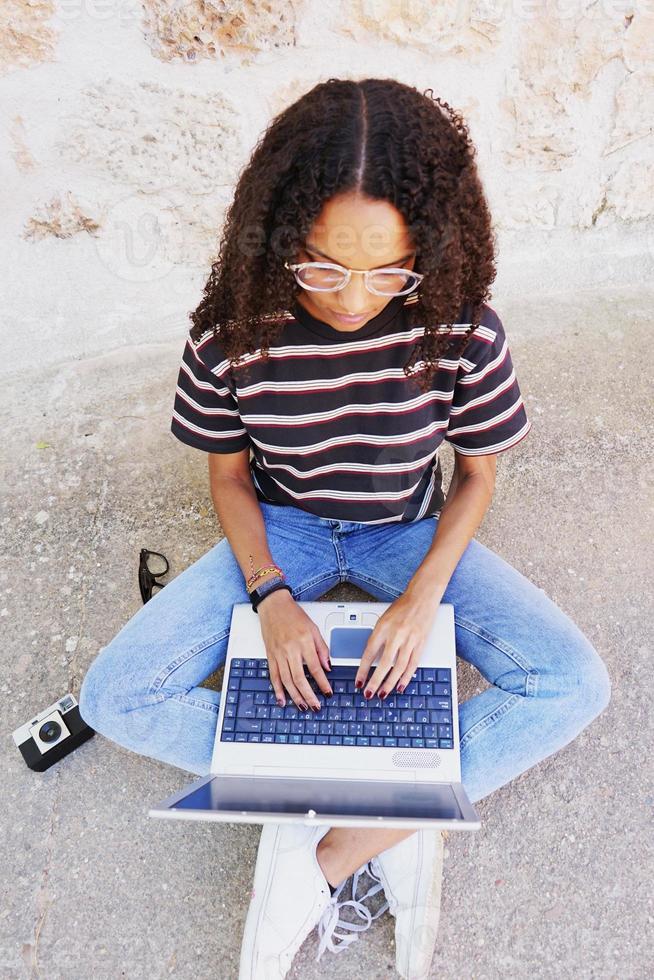 un portrait d'une jeune femme noire concentrée aux cheveux bouclés portant des lunettes, un jean et un t-shirt rayé, à côté de la technologie comme les smartphones et les appareils photo, assise par terre et travaillant ou faisant ses devoirs photo