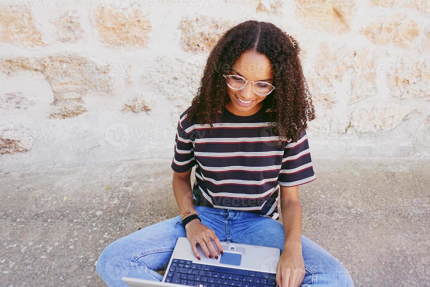 un portrait d'une jeune femme noire souriante et heureuse aux cheveux bouclés portant des lunettes, un jean et un t-shirt rayé, assise par terre et travaillant ou faisant ses devoirs photo