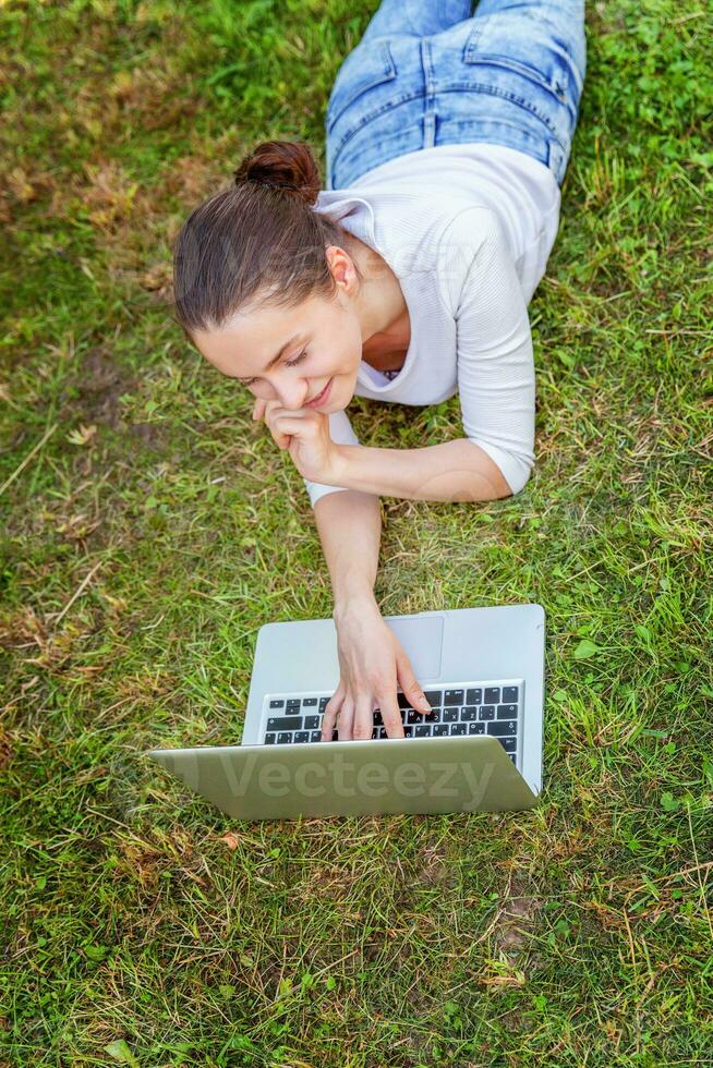 Jeune femme mensonge sur vert herbe pelouse dans ville parc travail sur portable PC ordinateur. free-lance affaires concept photo