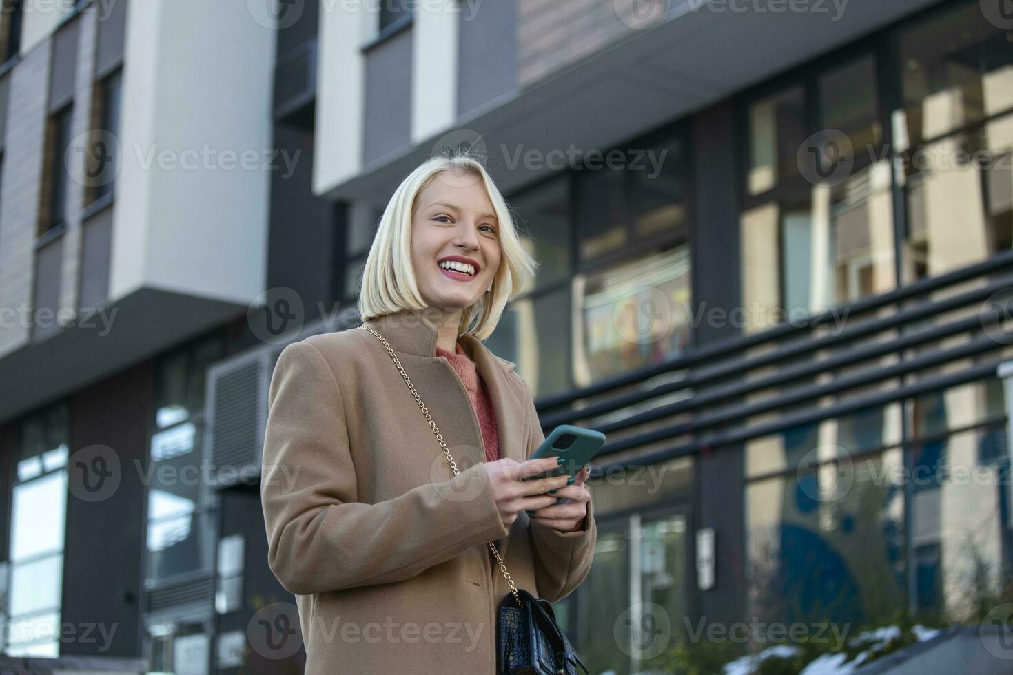 magnifique magnifique Jeune femme avec blond cheveux Messagerie sur le téléphone intelligent à le ville rue Contexte. jolie fille ayant intelligent téléphone conversation photo