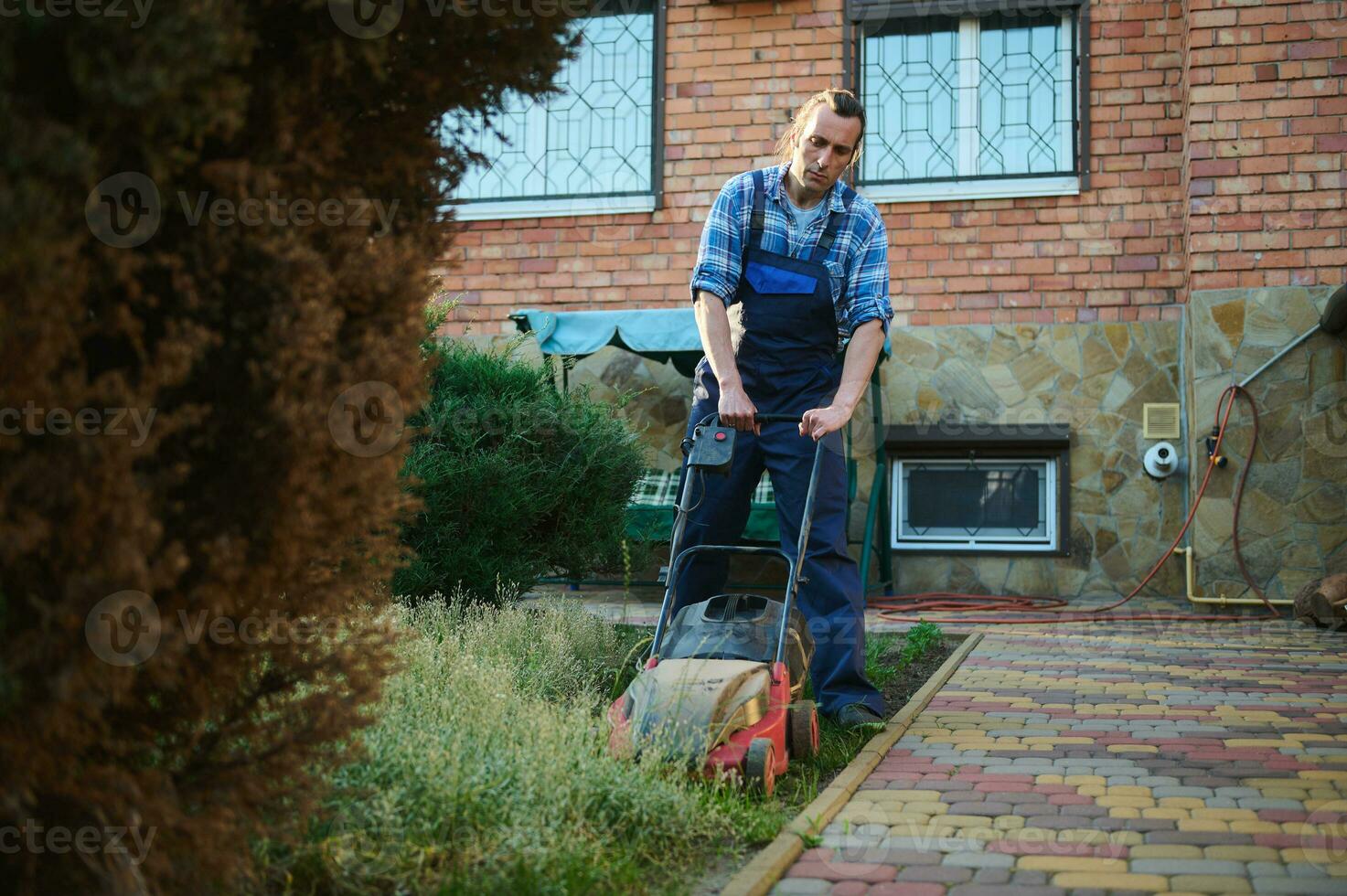 caucasien Beau ouvrier, Masculin jardinier paysagiste fauchage une Résidentiel pelouse avec un électrique pelouse tondeuse. photo