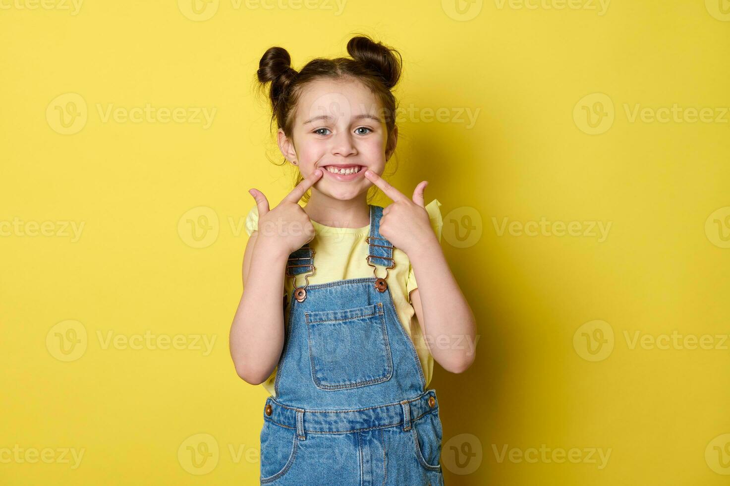 mignonne peu enfant fille dans décontractée denim porter, points sa les doigts à sa bouche, montrant une magnifique sourire, à la recherche à caméra photo