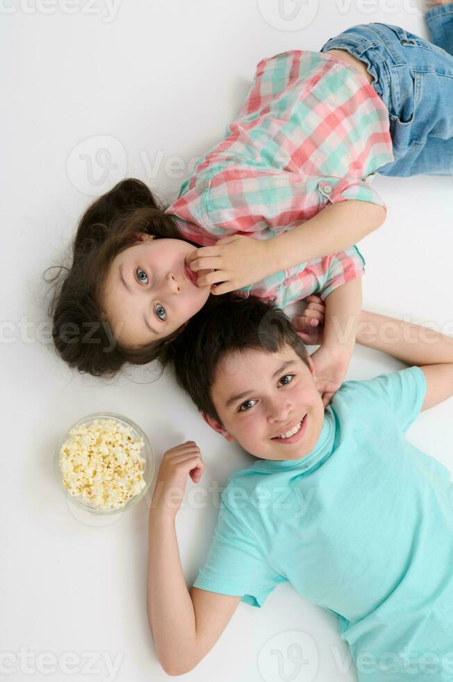 Haut vue content des gamins souriant à la recherche à caméra, mensonge sur leur dos sur blanc Contexte près une bol avec savoureux pop corn photo