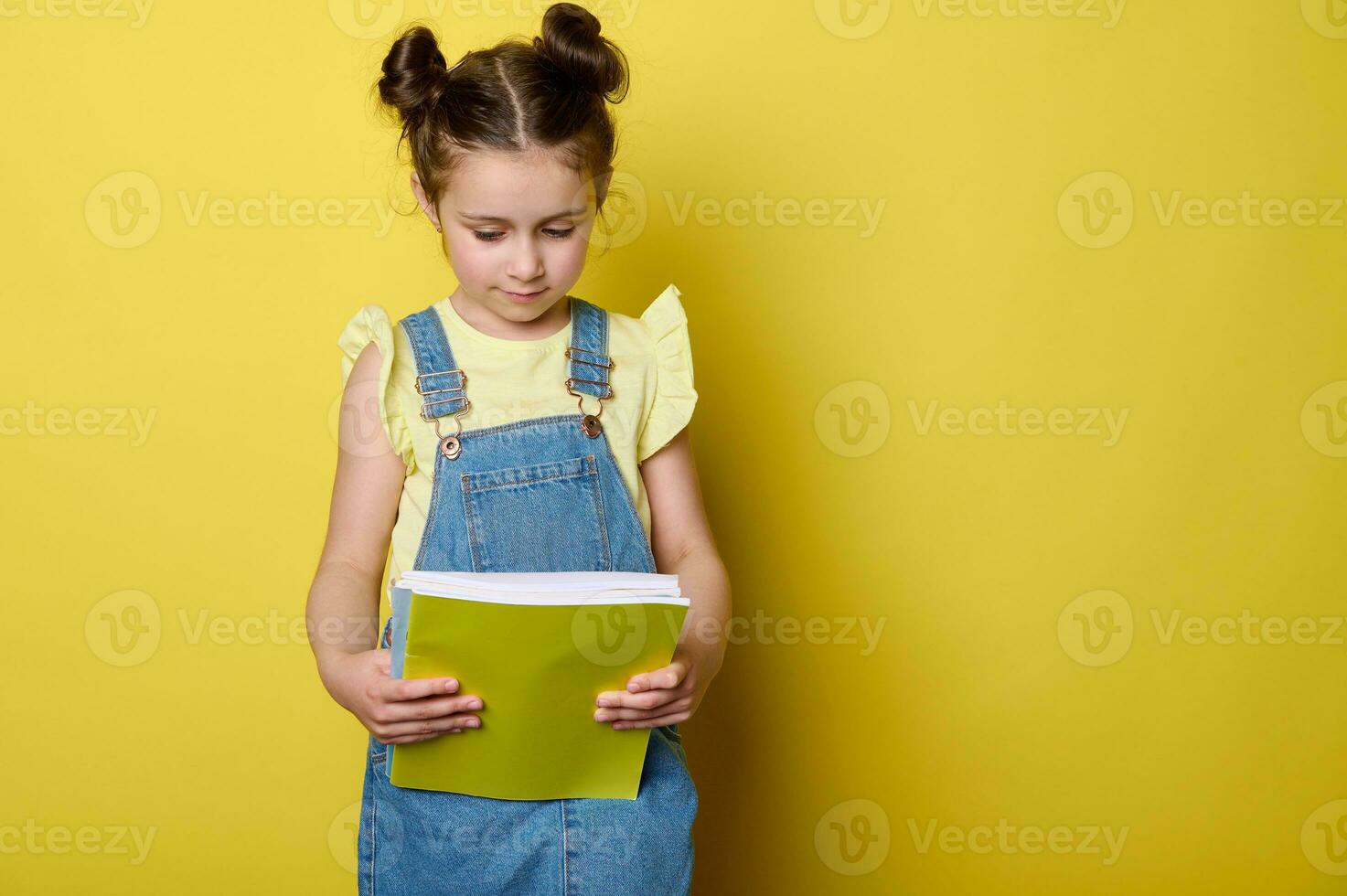 portrait de mignonne enfant fille dans Jaune T-shirt et bleu denim robe salopette, en portant cahier de texte isolé studio Contexte photo