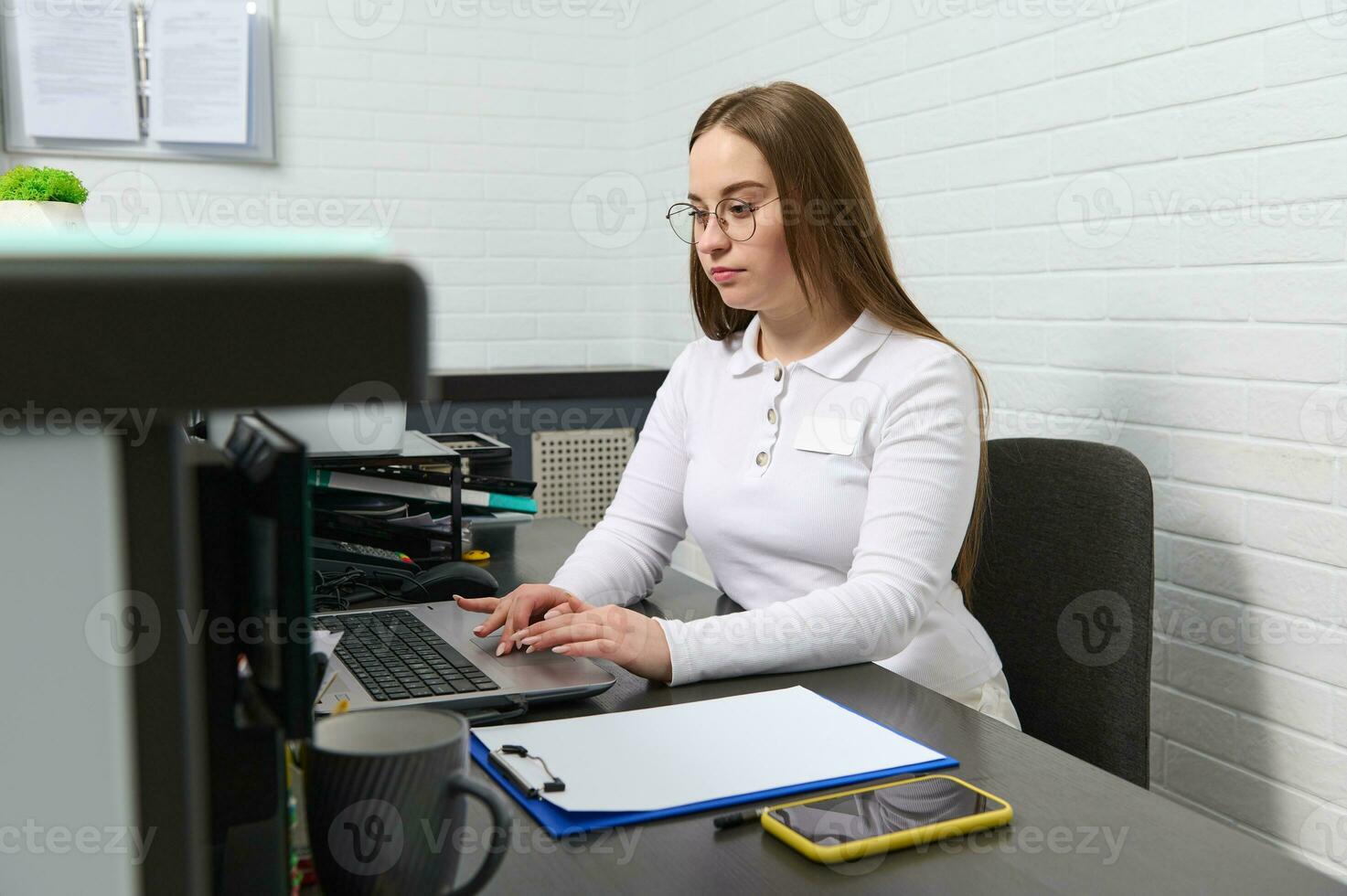 réceptionniste travaux sur ordinateur portable, séance derrière une accueil bureau dans moderne clinique, en gardant rendez-vous avec médical Personnel photo