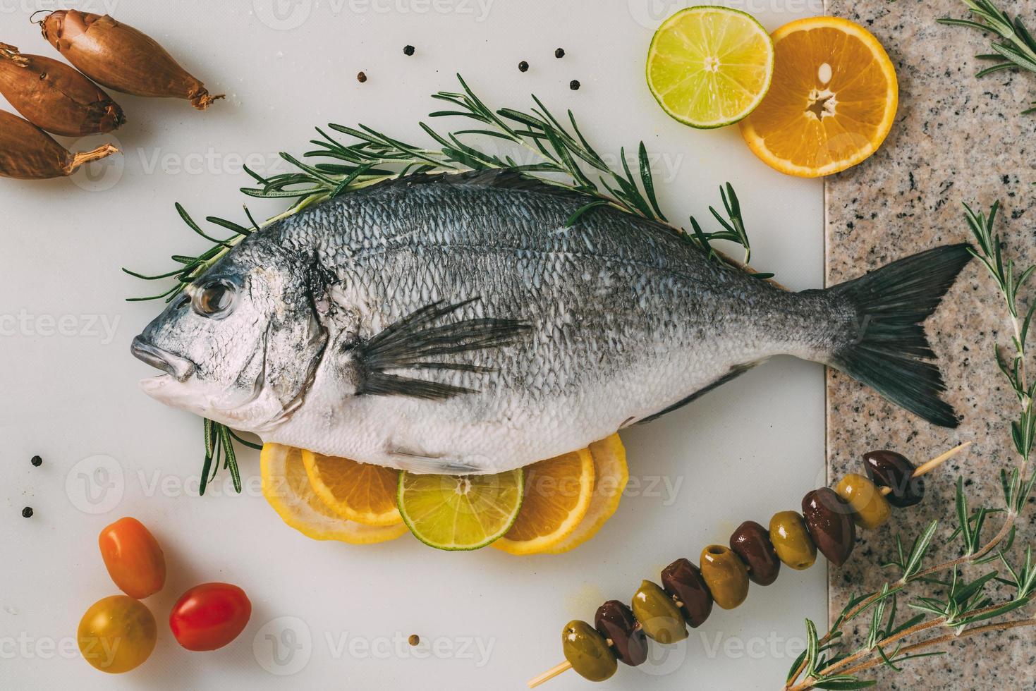 dorade de mer poisson sur table de cuisine avec romarin, citron, orange, olives, tomates, oignon et citron vert. orata frais, préparation de poisson dorade. photo