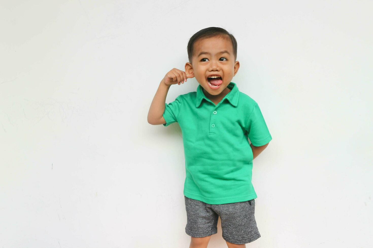 portrait de une mignonne peu asiatique garçon en portant le sien oreille tandis que collage le sien langue dehors, isolé sur le blanc Contexte photo