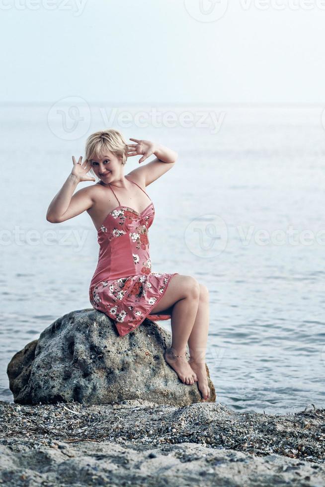 femme blonde heureuse dans une robe fleurie rougeâtre. femme assise et pose sur la grosse pierre au bord de la mer. photo