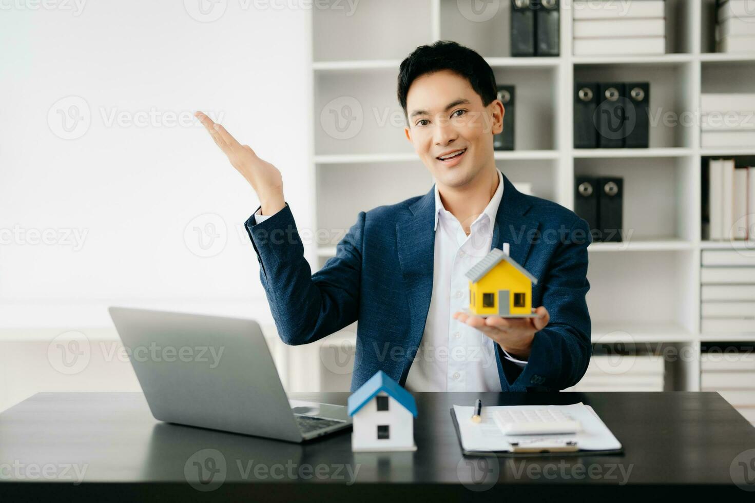 Jeune réel biens agent ouvrier travail avec portable et tablette à table dans moderne Bureau et petit maison à côté de il. photo