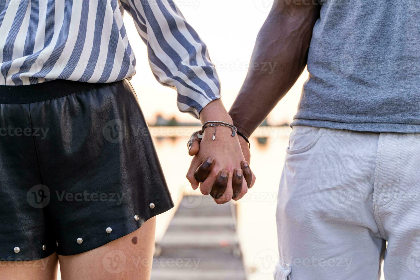 Jeune multiracial aimant couple en portant mains sur une quai de une rivière Port à le coucher du soleil photo