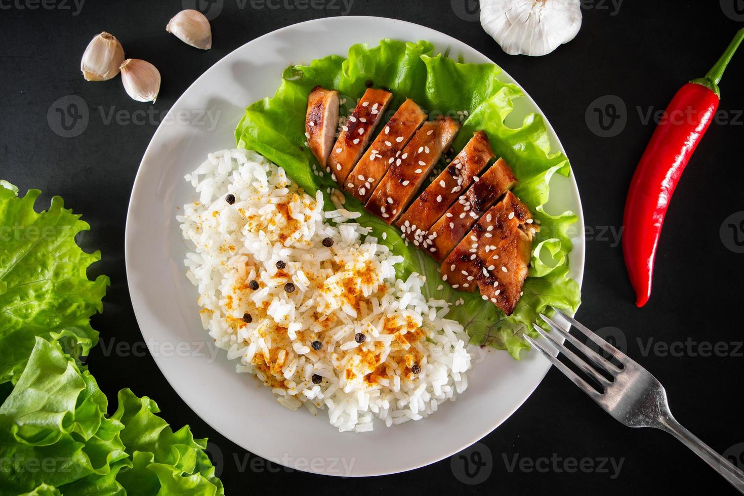 poulet teriyaki aux graines de sésame, laitue et riz sur une assiette blanche. plat avec une fourchette sur fond noir photo