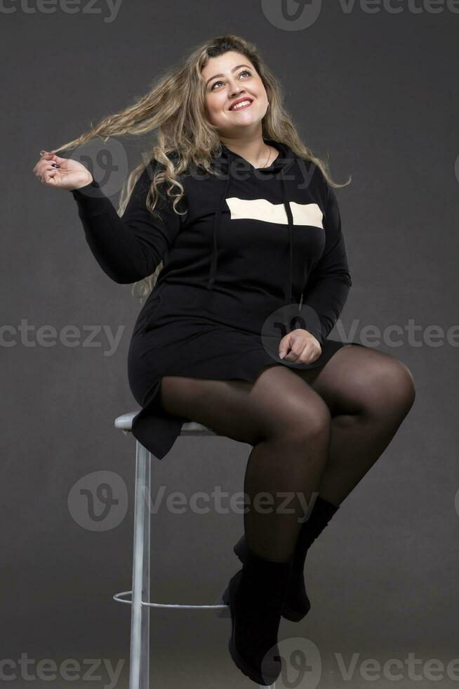 une magnifique graisse femme avec longue frisé cheveux est assis sur une chaise. photo