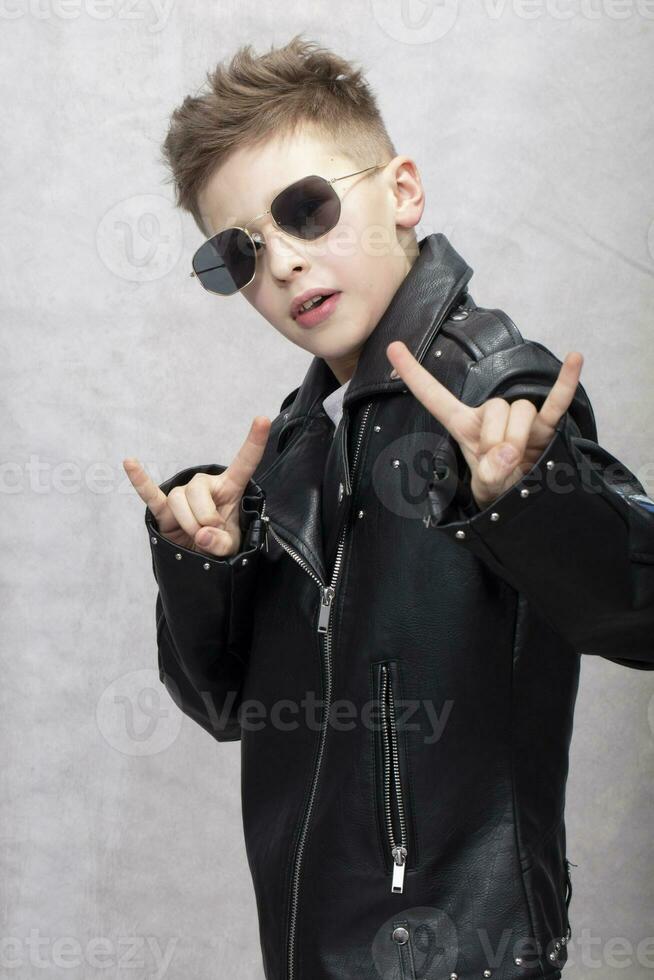 portrait de une lourd métal garçon dans des lunettes de soleil. mignonne adolescent dans une cuir veste Faire une Roche et rouleau signe photo