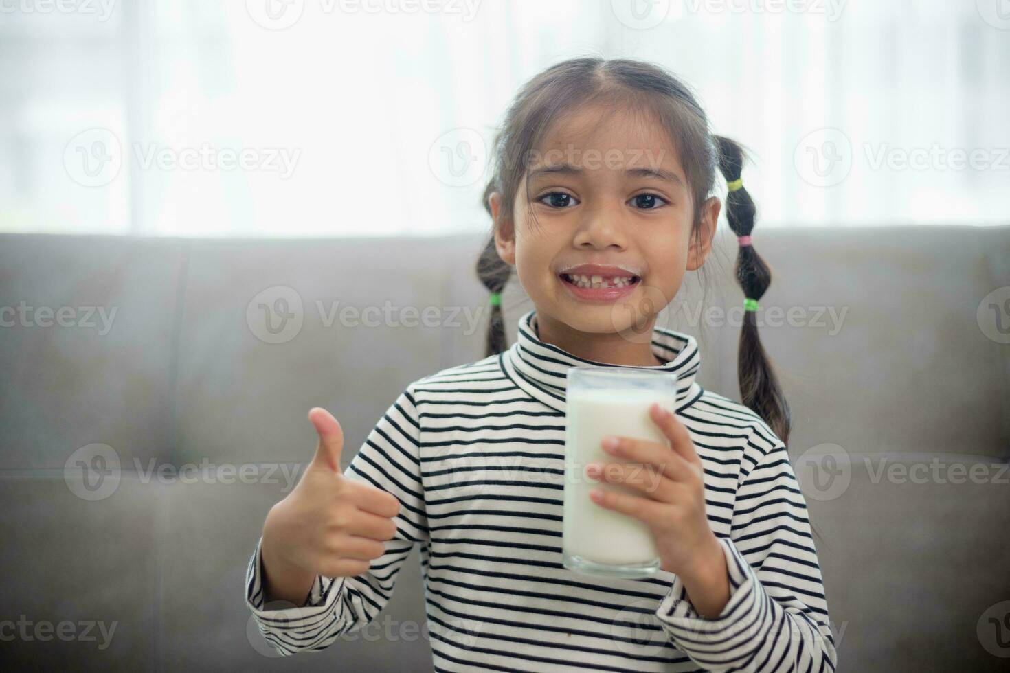 asiatique peu mignonne enfant en portant une tasse de Lait dans le maison. ressentir content et prendre plaisir en buvant lait. photo