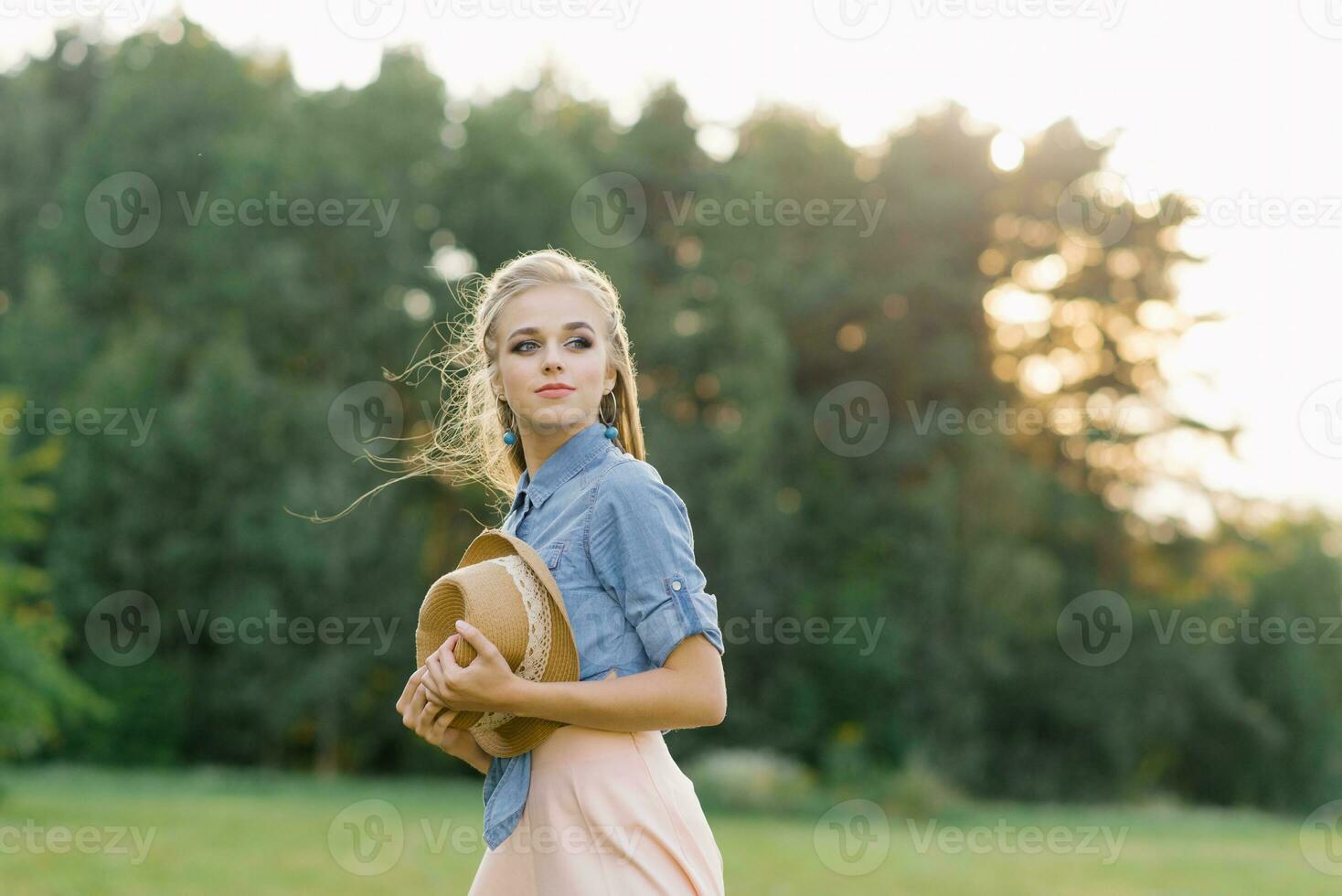 content femme avec longue cheveux avec une chapeau dans sa mains en marchant sur le vert herbe dans été photo