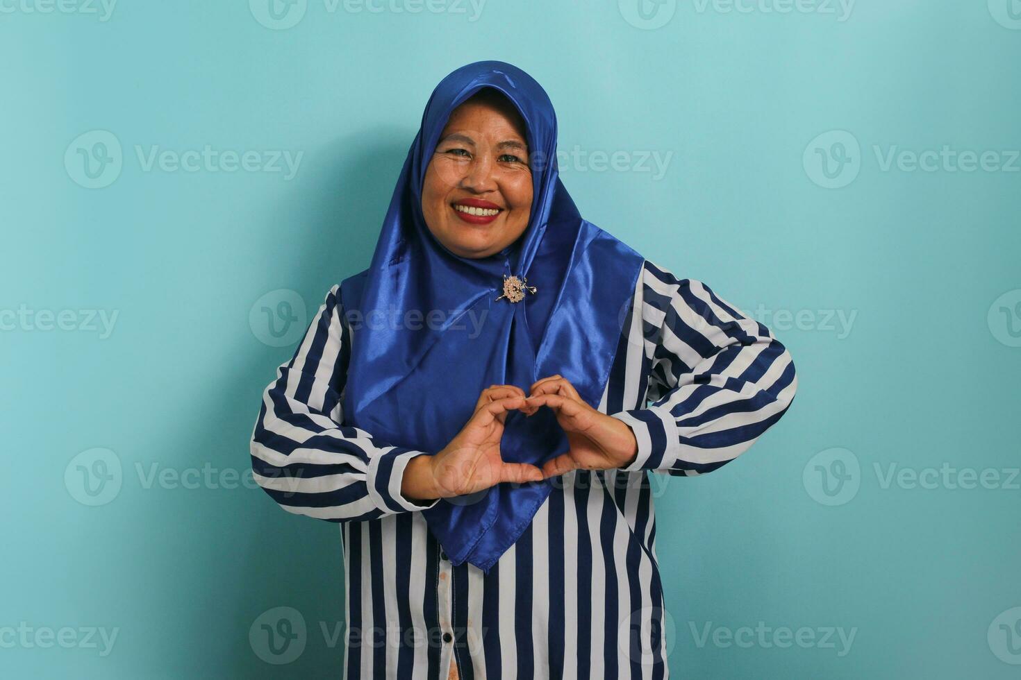 une souriant âge moyen asiatique femme, dans une bleu hijab et rayé chemise, fait du une cœur geste avec sa mains, rayonnant bonheur et se soucier, isolé sur bleu Contexte photo