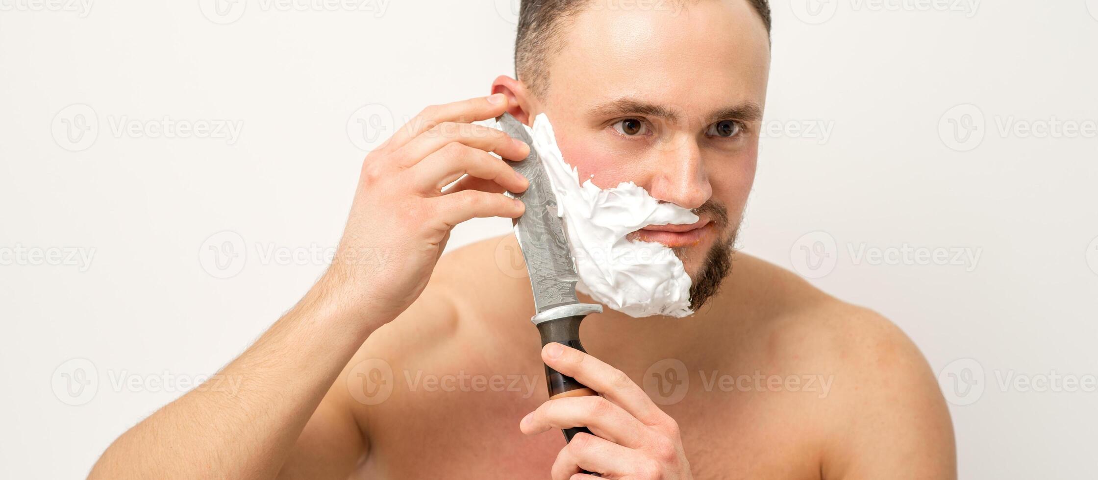 Jeune caucasien homme rasage barbe avec une gros couteau sur blanc Contexte. photo