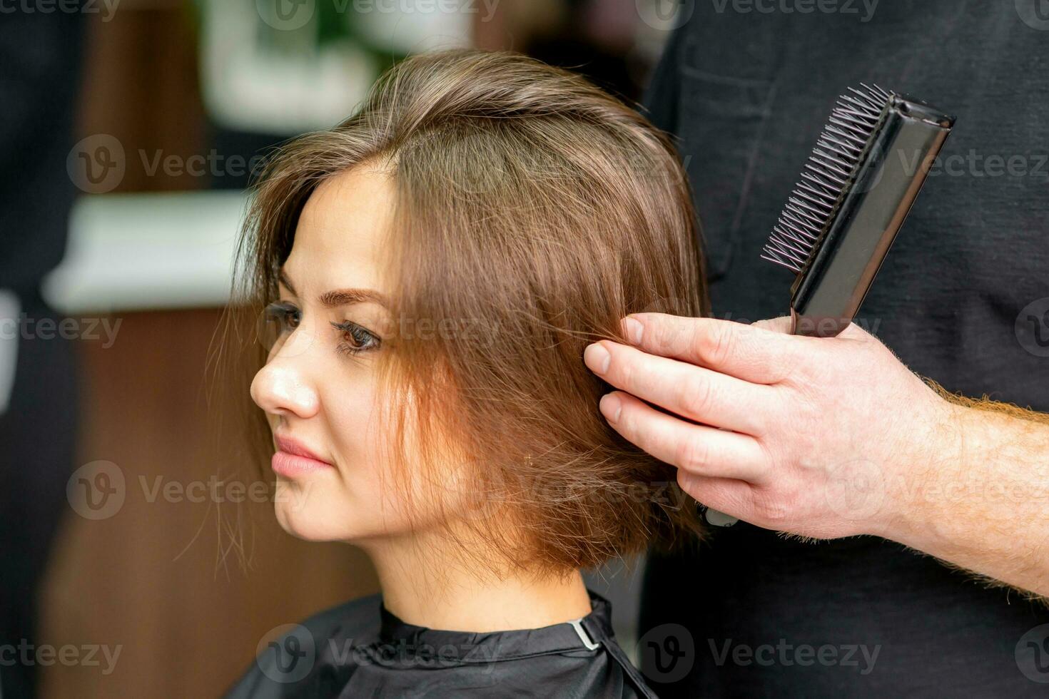 Masculin coiffeur travaux sur le coiffure de le Jeune caucasien brunette femme à une cheveux salon. photo