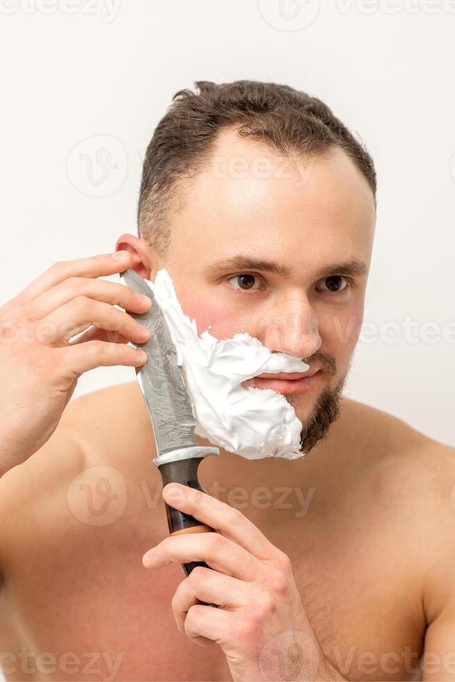 Jeune caucasien homme rasage barbe avec une gros couteau sur blanc Contexte. photo