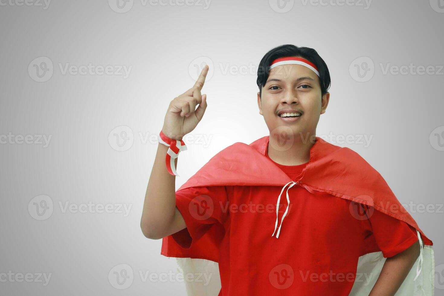 portrait de attrayant asiatique homme dans T-shirt avec rouge blanc ruban sur tête avec drapeau sur le sien épaule comme une manteau, montrer du doigt en haut le sien index et à la recherche à caméra. isolé image sur gris Contexte photo