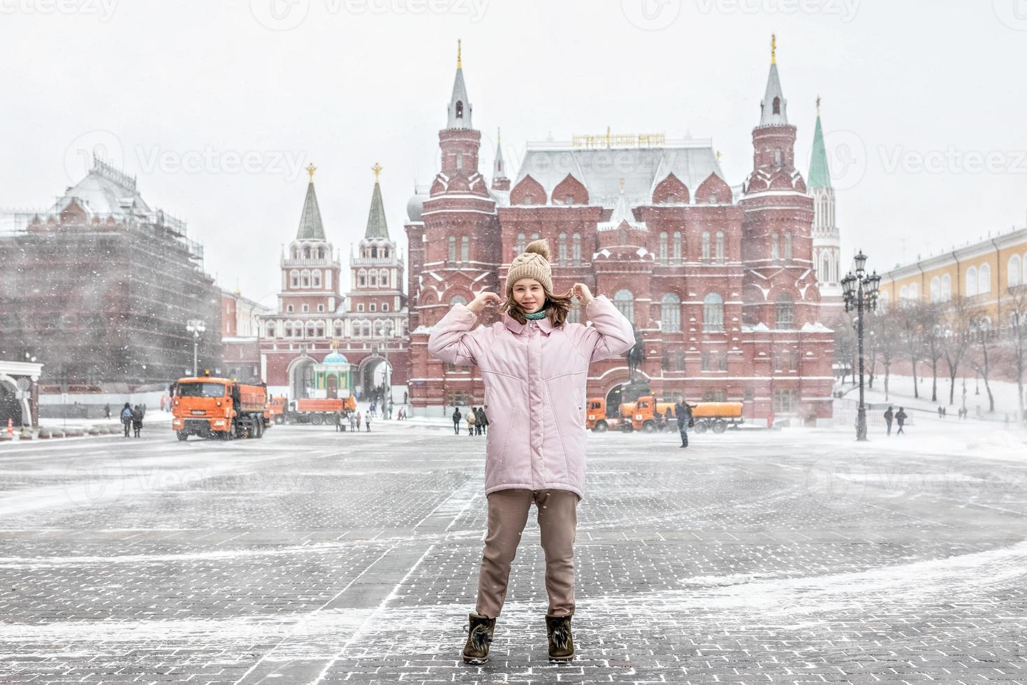 une belle jeune fille vêtue d'une veste rose se promène le long de la place manezhnaya à moscou lors d'une chute de neige et d'un blizzard. les souffleuses à neige travaillent en arrière-plan. photo