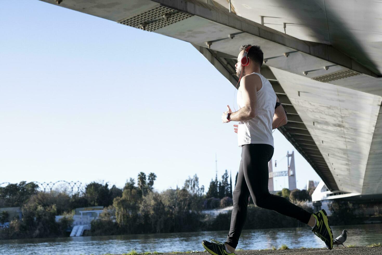 Jeune homme fonctionnement dans une blanc chemise à travers une pont. il est écoute à la musique et il est eu certains casques sur. photo