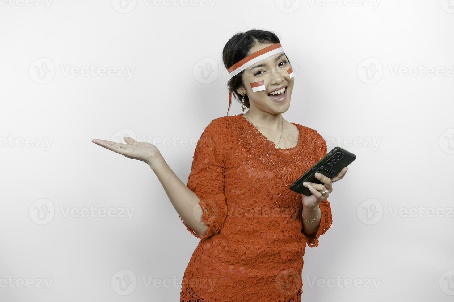 une content asiatique femme portant rouge kebaya et Bandeau, en portant sa téléphone, et montrer du doigt copie espace à côté de son, isolé par blanc Contexte. l'indonésie indépendance journée photo
