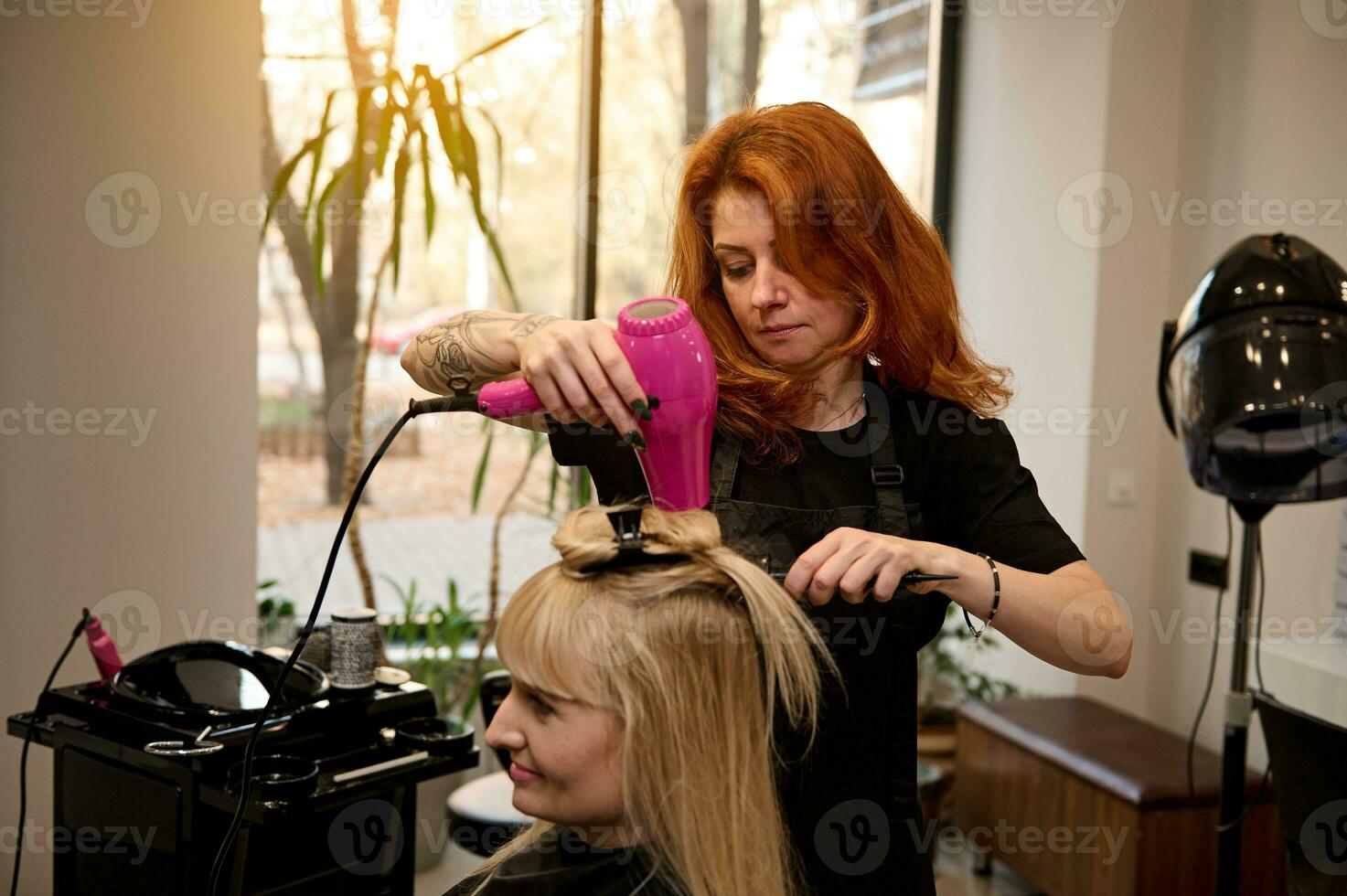 à la mode coiffeur avec tatoué bras en utilisant une rond peigne et cheveux séchoir se redresse blond cheveux à une client de une beauté salon, performant sa une élégant coiffure. beauté industrie concept photo