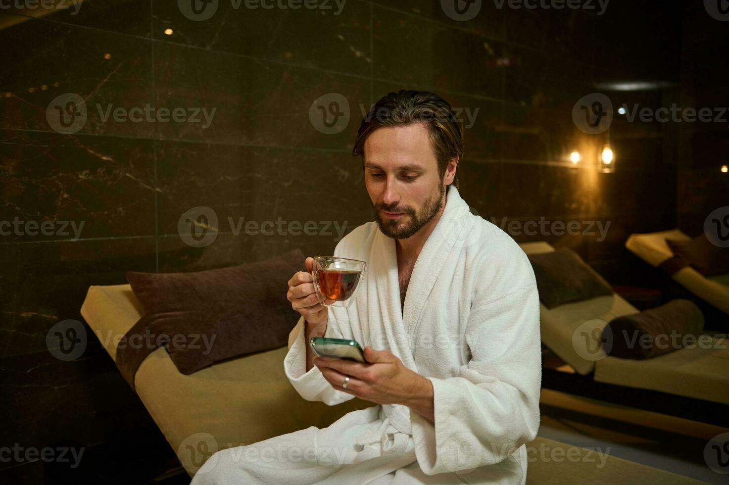 Beau attrayant caucasien homme dans peignoir de bain relaxant, en utilisant mobile téléphone, navigation la toile des sites et en buvant à base de plantes thé, séance sur une cabriolet salon par jacuzzi dans luxueux ayurvédique bien-être spa recours photo