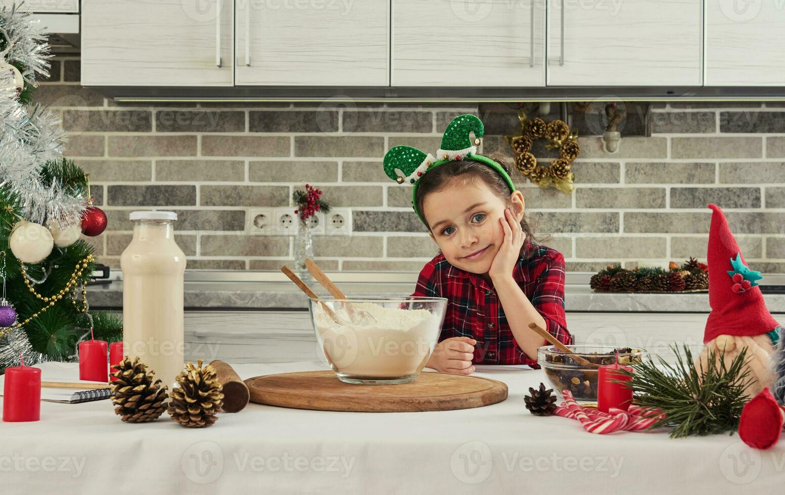 adorable caucasien peu fille dans rouge robe et elfe cerceau sur sa tête regards à caméra séance à une table avec Ingrédients pour fabrication sucré pâte pour Noël gâteau et Fait main scandinave gnomes photo