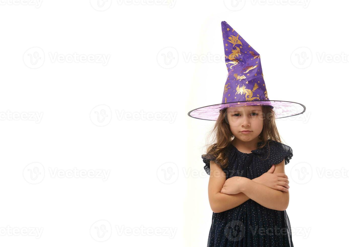 portrait de mignonne peu sorcière en colère dérangé fille portant une sorcier chapeau et habillé dans élégant carnaval robe, à la recherche à caméra posant avec franchi bras contre blanc arrière-plan, copie espace, Halloween photo