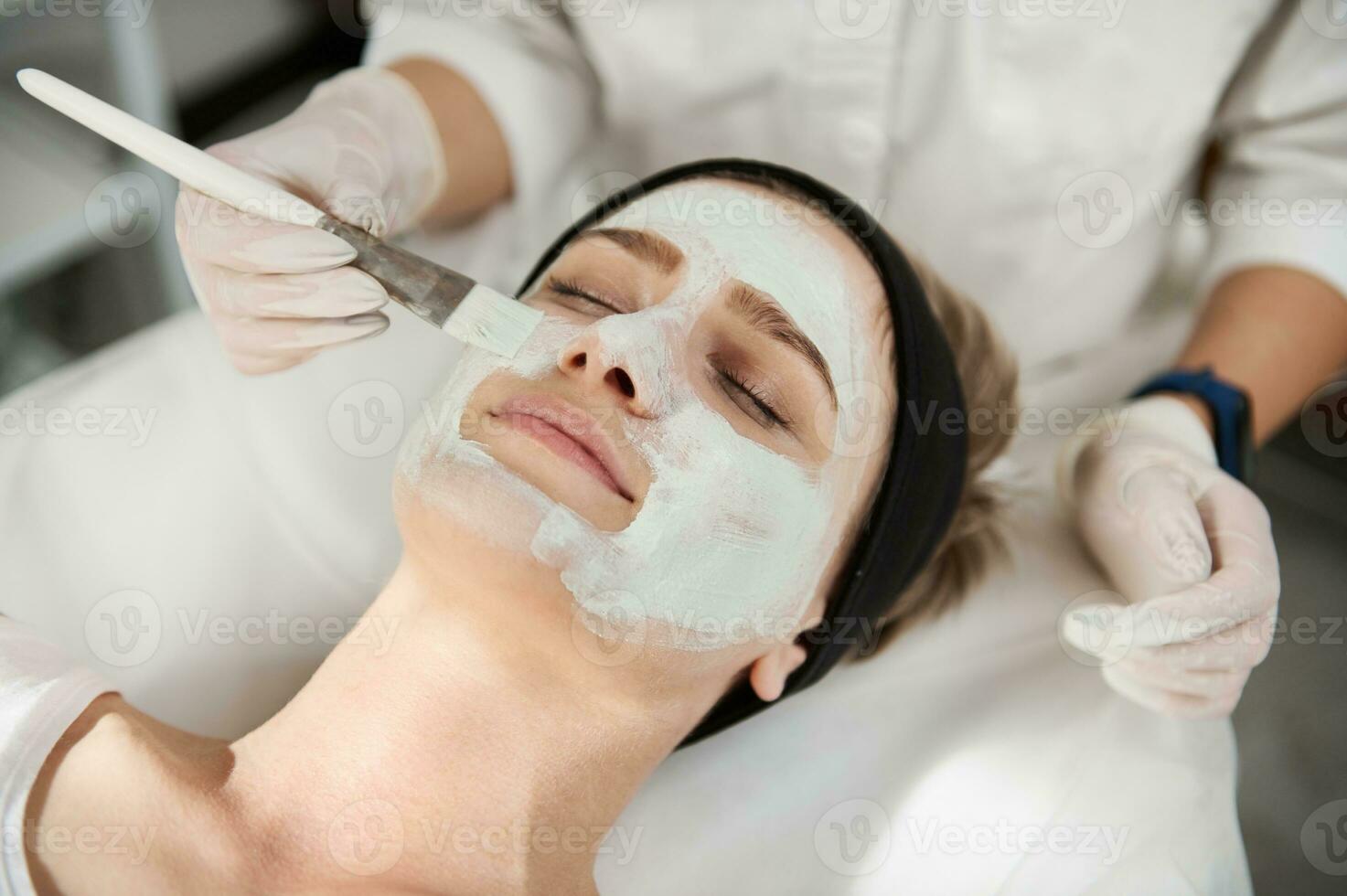 fermer portrait de une esthéticienne main appliquant une beauté masque sur le visage de une Jeune femme avec parfait nettoyer peau avec une brosse. professionnel peau se soucier concept dans moderne beauté spa salon photo