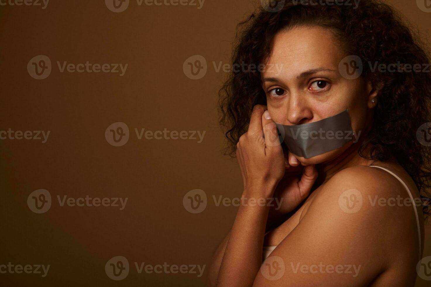 épuisé effrayé femme avec scellé bouche regards désespérément à le caméra, permanent de côté à une foncé beige Contexte avec espace pour texte. social concept de fin la violence contre femmes photo