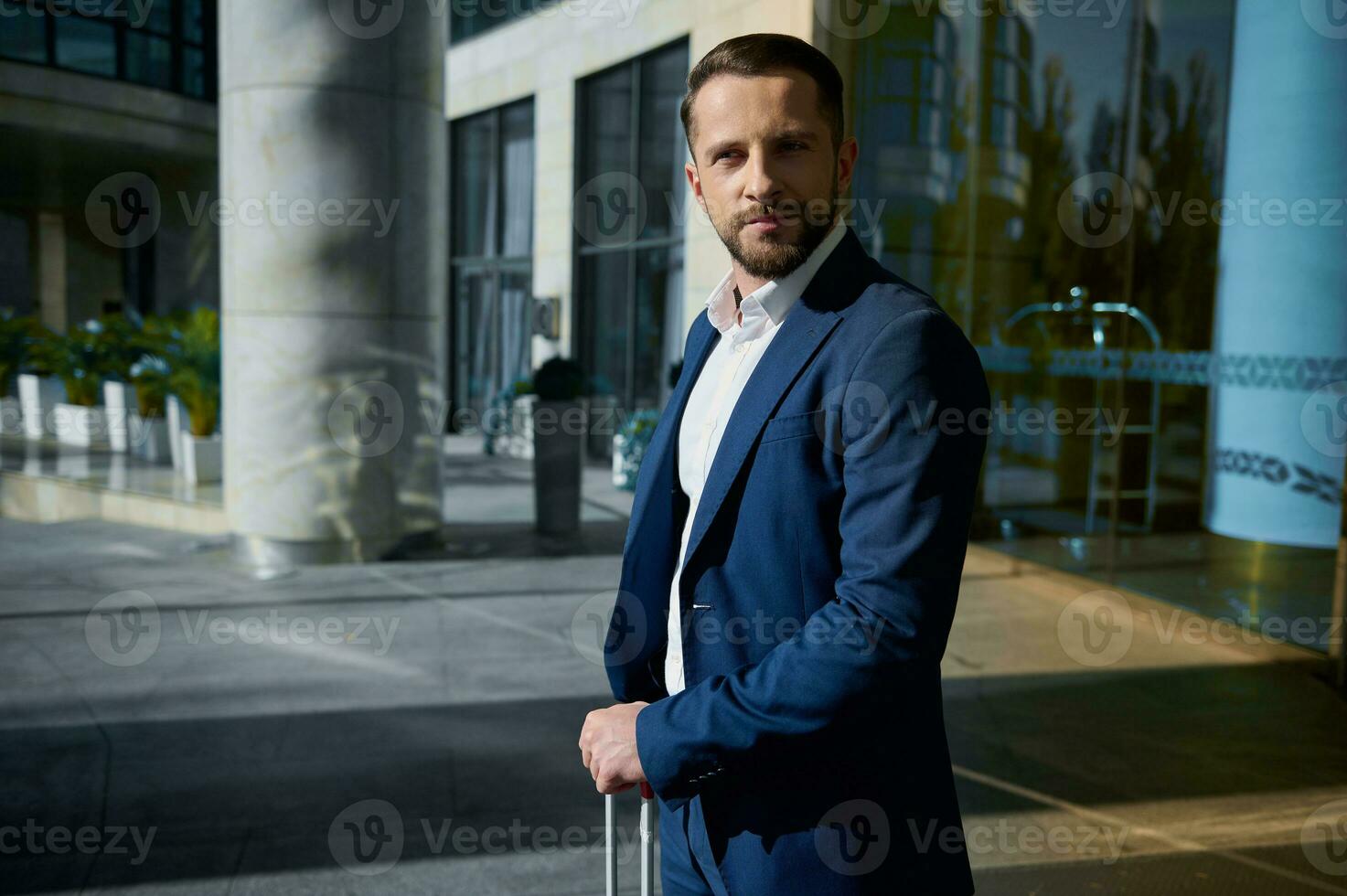 taille longueur portrait de une Beau réussi homme d'affaire développeur entrepreneur dans affaires costume avec une bagage permanent Extérieur sur une entreprise moderne gratte-ciel bâtiment verre entrée Contexte photo