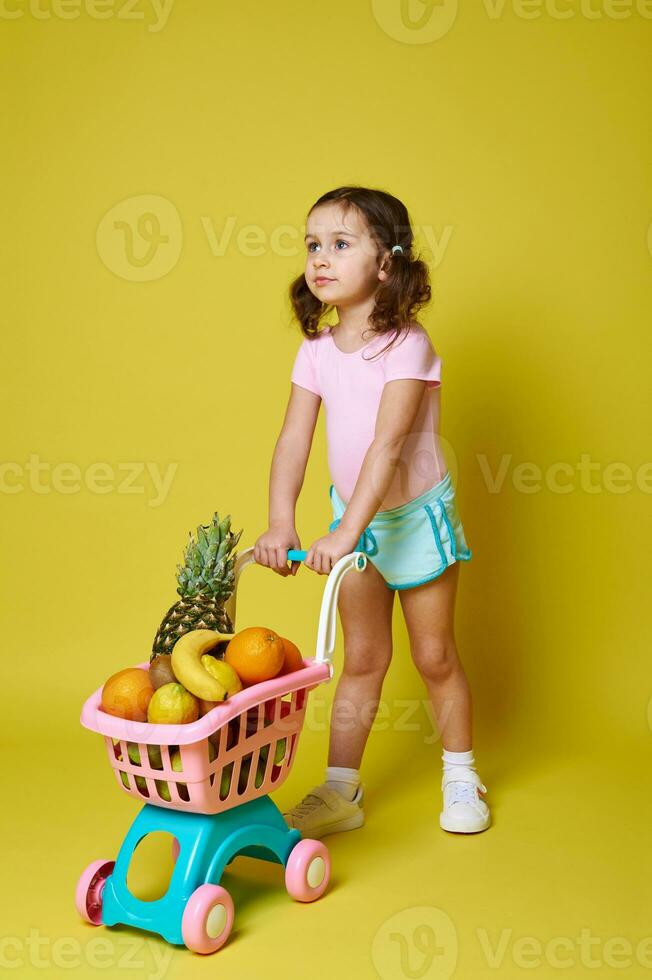 mignonne peu fille dans rose le maillot de corps et bleu été short Rouleaux une achats Chariot plein de des fruits. Jaune Contexte. copie espace pour La publicité photo