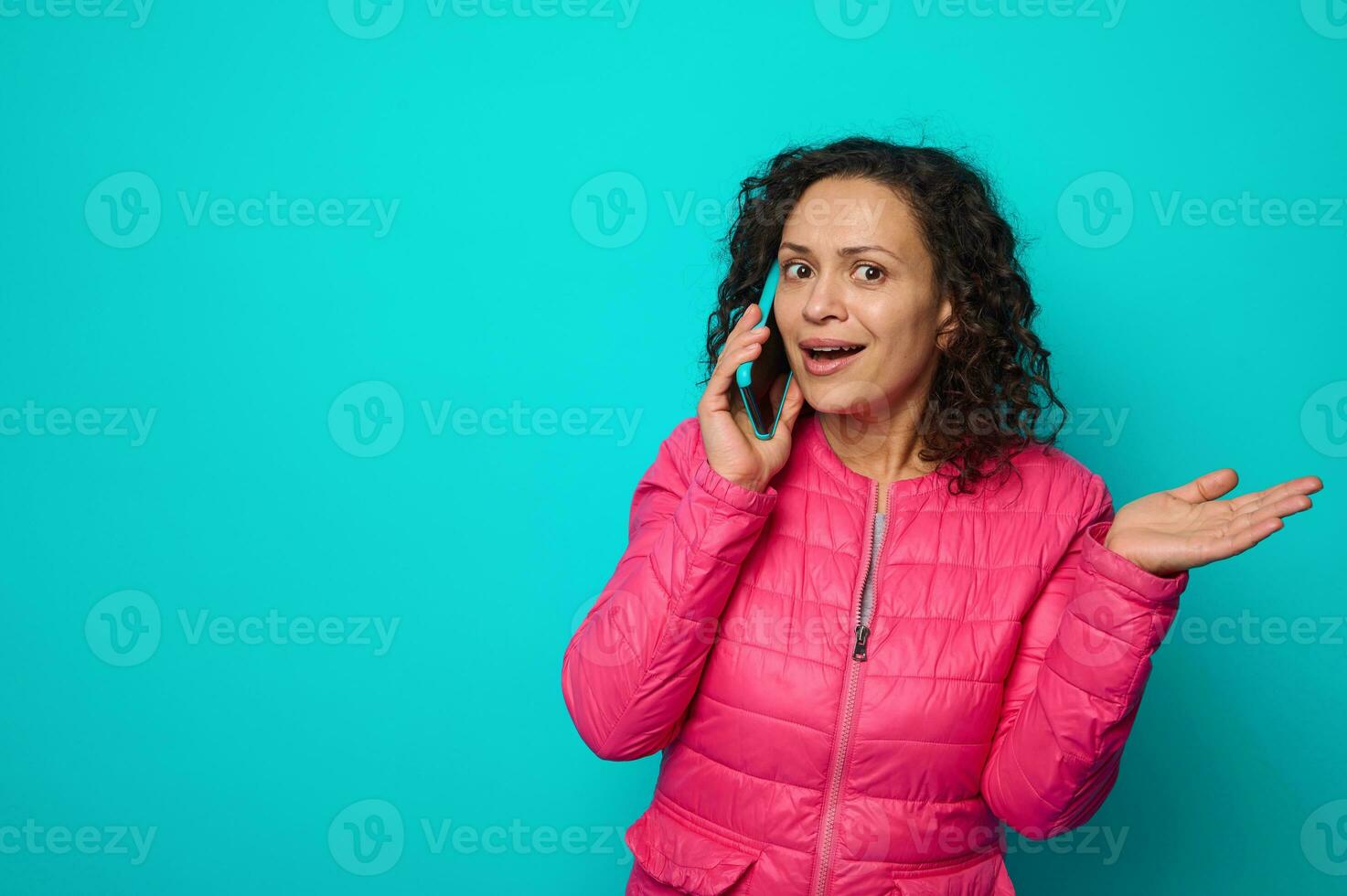 surpris sous le choc émotif frisé aux cheveux magnifique femme dans brillant rose veste pourparlers sur mobile téléphone, et gestes avec sa main paume en haut, isolé sur bleu coloré Contexte avec copie espace photo