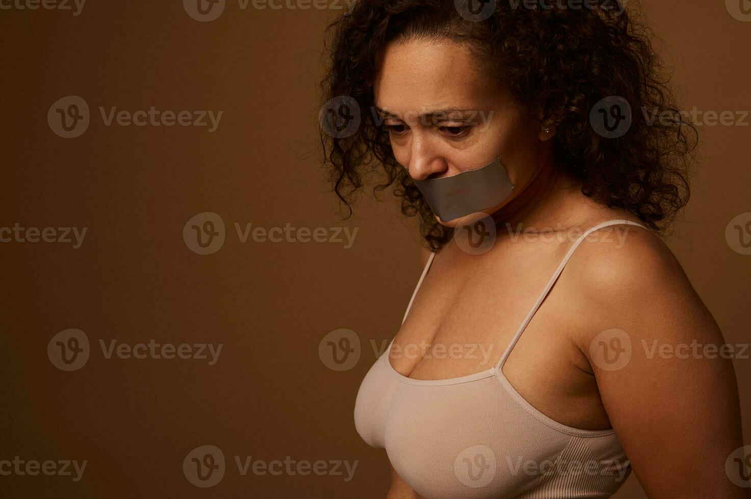 épuisé effrayé femme avec scellé bouche regards désespérément bas, permanent Trois quarts contre une foncé beige Contexte avec espace pour texte. social concept de fin la violence contre femmes photo