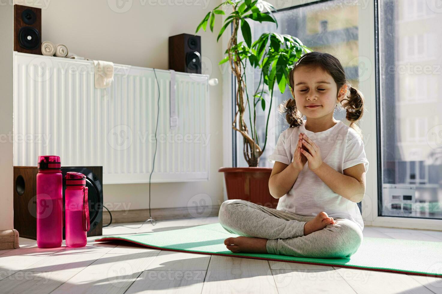 magnifique peu fille séance sur une aptitude tapis et pratiquant yoga dans lotus position contre le Contexte de grand les fenêtres à Accueil photo