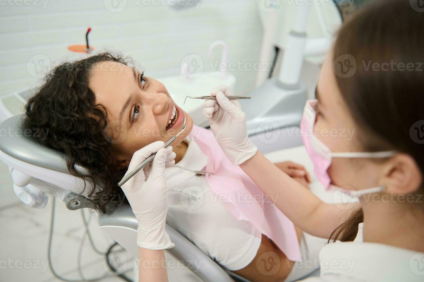 magnifique femme dans dentiste chaise pendant dentaire vérification dans dentisterie clinique sur flou premier plan de dentaire hygiéniste en portant inoxydable acier dentaire instruments et examiner les patients oral cavité photo